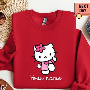 Puede incluir: Una sudadera roja con un personaje de dibujos animados con un vestido y una flor rosas. El texto "Your name" est&aacute; impreso en blanco. La sudadera tiene cuello redondo y mangas largas.