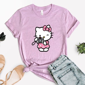 Pode incluir: Uma camiseta rosa com uma imagem de desenho animado do Hello Kitty segurando uma c&acirc;mera. A camiseta &eacute; feita de um material macio e confort&aacute;vel e tem um caimento relaxado.