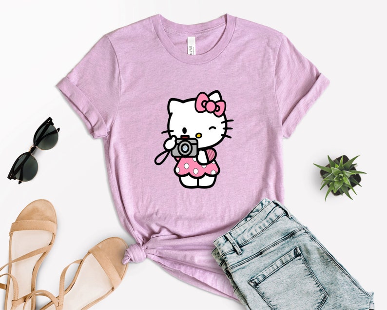 Pode incluir: Uma camiseta rosa com uma imagem de desenho animado do Hello Kitty segurando uma c&acirc;mera. A camiseta &eacute; feita de um material macio e confort&aacute;vel e tem um caimento relaxado.
