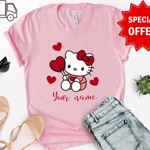 May include: Pink t-shirt with a cartoon image of Hello Kitty holding a red heart-shaped lollipop. There are red hearts scattered around Hello Kitty. The text "Your name" is printed below Hello Kitty.