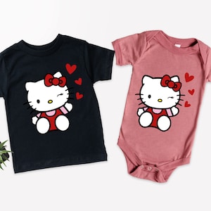 Op de afbeelding: Twee T-shirts met een cartoon van Hello Kitty met rode hartjes. Het shirt aan de linkerkant is zwart en het shirt aan de rechterkant is roze.
