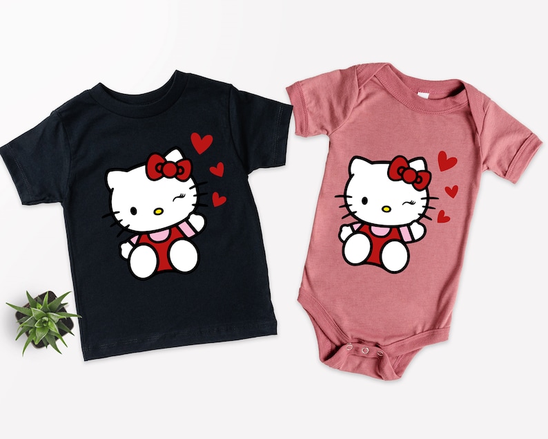Op de afbeelding: Twee T-shirts met een cartoon van Hello Kitty met rode hartjes. Het shirt aan de linkerkant is zwart en het shirt aan de rechterkant is roze.
