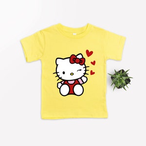 Op de afbeelding: Geel T-shirt met een cartoon afbeelding van Hello Kitty die knipoogt en een kusje blaast. De afbeelding is omringd door rode hartjes.