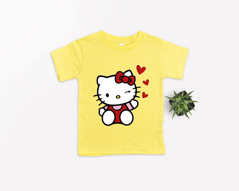 Op de afbeelding: Geel T-shirt met een cartoon afbeelding van Hello Kitty die knipoogt en een kusje blaast. De afbeelding is omringd door rode hartjes.