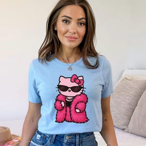 Puede incluir: Camiseta azul claro con un gato de dibujos animados que lleva gafas de sol y un abrigo de piel rosa. El gato tiene un lazo rosa y un collar de perlas. La camiseta es de cuello redondo y est&aacute; hecha de un material suave.