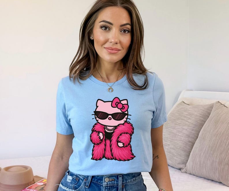 Puede incluir: Camiseta azul claro con un gato de dibujos animados que lleva gafas de sol y un abrigo de piel rosa. El gato tiene un lazo rosa y un collar de perlas. La camiseta es de cuello redondo y est&aacute; hecha de un material suave.