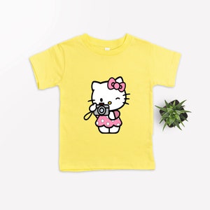 Pode incluir: Uma camiseta amarela com uma imagem de desenho animado do Hello Kitty segurando uma c&acirc;mera. Hello Kitty est&aacute; vestindo um vestido rosa com bolinhas brancas e um la&ccedil;o rosa.