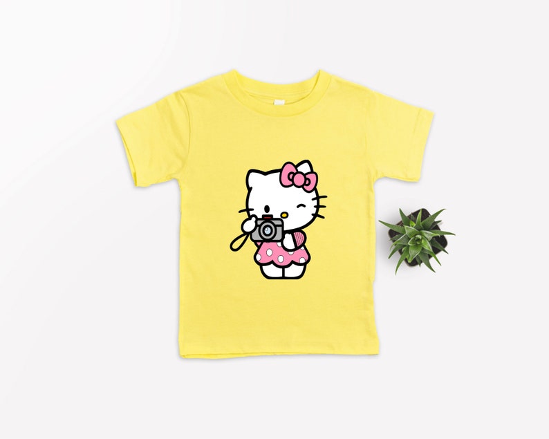 Pode incluir: Uma camiseta amarela com uma imagem de desenho animado do Hello Kitty segurando uma c&acirc;mera. Hello Kitty est&aacute; vestindo um vestido rosa com bolinhas brancas e um la&ccedil;o rosa.