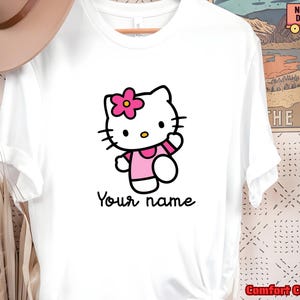 May include: White t-shirt featuring a cartoon image of Hello Kitty wearing a pink dress and a flower in her hair. The text "Your name" is printed below the character in a handwritten style font.