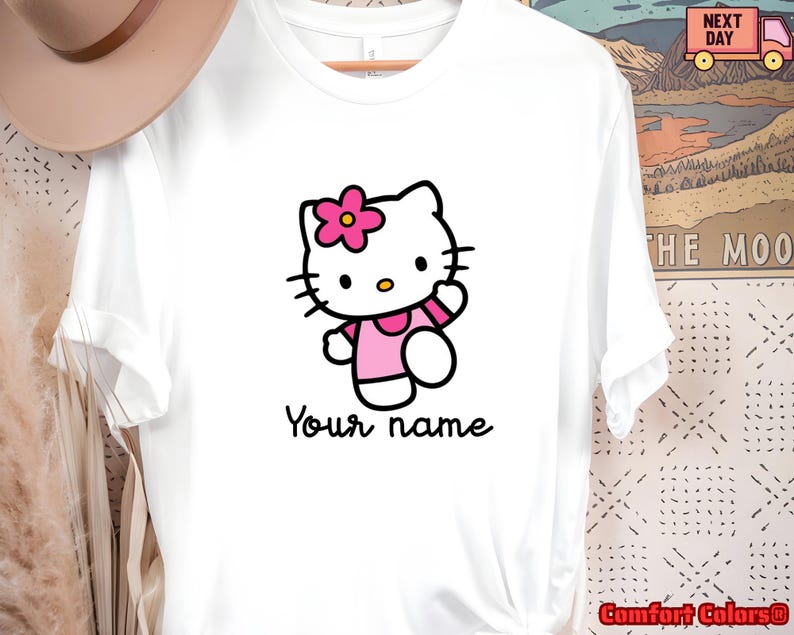 May include: White t-shirt featuring a cartoon image of Hello Kitty wearing a pink dress and a flower in her hair. The text "Your name" is printed below the character in a handwritten style font.