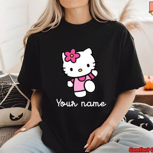 May include: Black t-shirt featuring a cartoon character with a pink flower, wearing a pink dress. The text "Your name" is written in white script below the character. The shirt is a Comfort Colors brand.