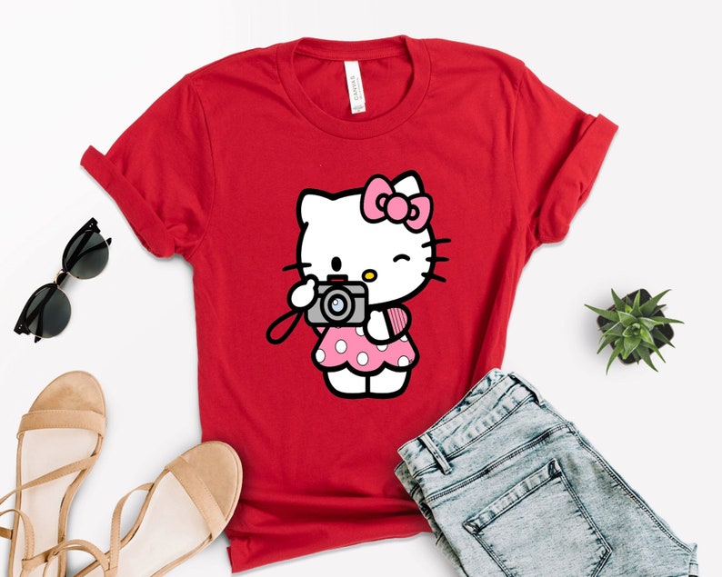 Pode incluir: Camiseta vermelha com um gr&aacute;fico branco de Hello Kitty. Hello Kitty est&aacute; segurando uma c&acirc;mera rosa e preta e vestindo um vestido rosa com bolinhas brancas.
