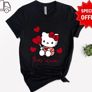 May include: Black t-shirt with a white Hello Kitty character holding a red heart lollipop. The character is surrounded by red hearts. The text "Your name" is printed below the character.
