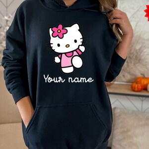 Puede incluir: Una sudadera con capucha negra con un personaje de dibujos animados con una flor rosa, que lleva un vestido rosa y saluda. El texto "Your name" est&aacute; impreso en blanco debajo. Una pegatina roja de "Special Offer" es visible.