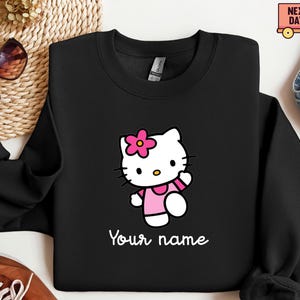 Puede incluir: Sudadera negra con un personaje de gato de dibujos animados con una flor rosa, un vestido y brazaletes. El gato est&aacute; caminando y el texto "Your name" est&aacute; escrito en blanco debajo. La sudadera tiene cuello redondo.