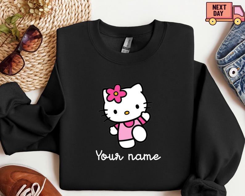 Puede incluir: Sudadera negra con un personaje de gato de dibujos animados con una flor rosa, un vestido y brazaletes. El gato est&aacute; caminando y el texto "Your name" est&aacute; escrito en blanco debajo. La sudadera tiene cuello redondo.
