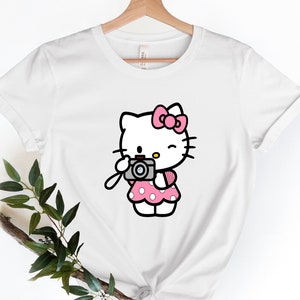 Pode incluir: Camiseta branca com um gr&aacute;fico rosa de Hello Kitty segurando uma c&acirc;mera. Hello Kitty est&aacute; vestindo um vestido rosa de bolinhas e um la&ccedil;o rosa.