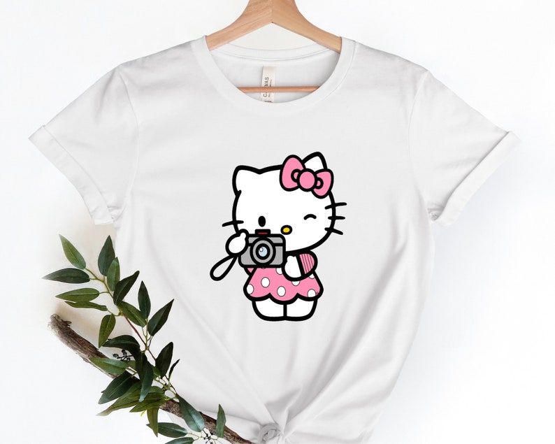 Pode incluir: Camiseta branca com um gr&aacute;fico rosa de Hello Kitty segurando uma c&acirc;mera. Hello Kitty est&aacute; vestindo um vestido rosa de bolinhas e um la&ccedil;o rosa.