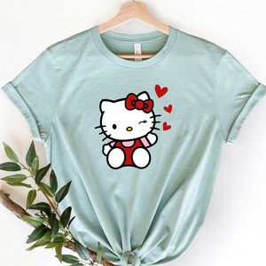 Op de afbeelding: Een lichtblauw t-shirt met een cartoon afbeelding van Hello Kitty die knipoogt en rode hartjes blaast.
