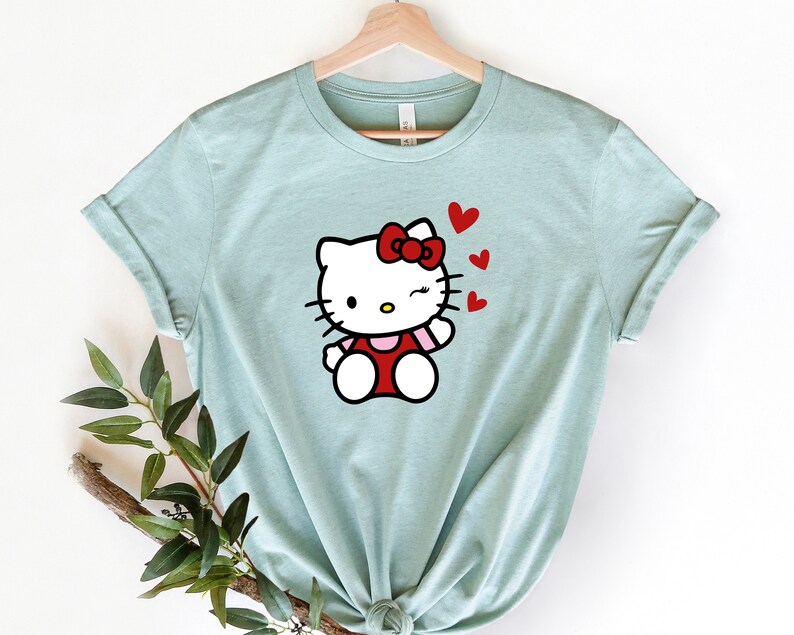 Op de afbeelding: Een lichtblauw t-shirt met een cartoon afbeelding van Hello Kitty die knipoogt en rode hartjes blaast.