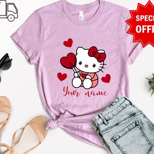 May include: Pink t-shirt with a cartoon image of Hello Kitty holding a red lollipop and surrounded by red hearts. The text "Your name" is printed below the image.