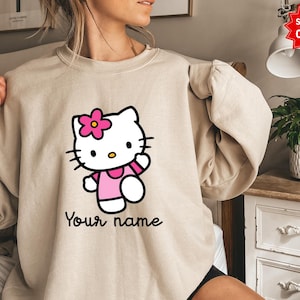 Puede incluir: Sudadera beige con un gr&aacute;fico de Hello Kitty. El gr&aacute;fico incluye una flor rosa, un vestido y un brazo. El texto "Your name" est&aacute; escrito en negro debajo. La sudadera es de estilo casual.