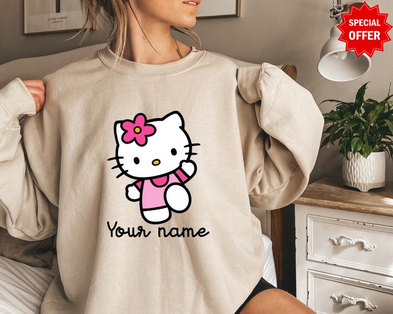 Puede incluir: Sudadera beige con un gr&aacute;fico de Hello Kitty. El gr&aacute;fico incluye una flor rosa, un vestido y un brazo. El texto "Your name" est&aacute; escrito en negro debajo. La sudadera es de estilo casual.