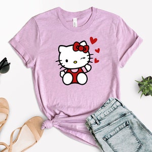 Op de afbeelding: Een roze T-shirt met een cartoon afbeelding van Hello Kitty die knipoogt en een kusje blaast. De afbeelding is omringd door rode hartjes.