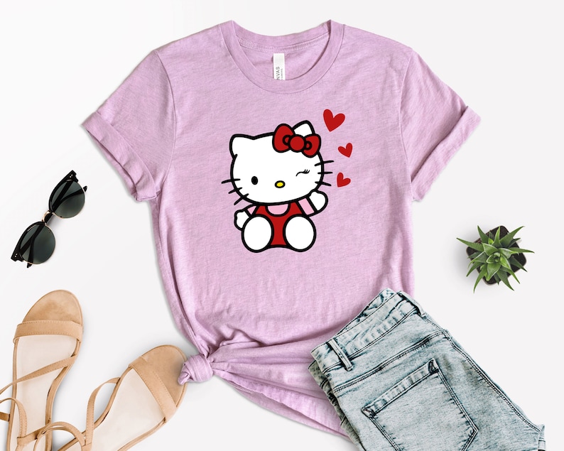 Op de afbeelding: Een roze T-shirt met een cartoon afbeelding van Hello Kitty die knipoogt en een kusje blaast. De afbeelding is omringd door rode hartjes.