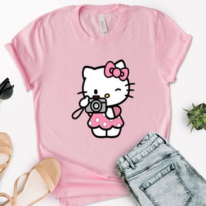 Pode incluir: Camiseta rosa com uma imagem de desenho animado do Hello Kitty segurando uma c&acirc;mera. Hello Kitty est&aacute; vestindo um vestido rosa com bolinhas brancas e um la&ccedil;o rosa.