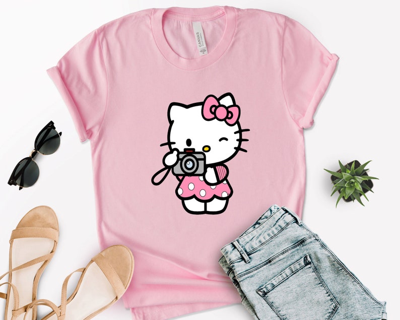 Pode incluir: Camiseta rosa com uma imagem de desenho animado do Hello Kitty segurando uma c&acirc;mera. Hello Kitty est&aacute; vestindo um vestido rosa com bolinhas brancas e um la&ccedil;o rosa.