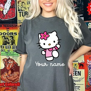 May include: A charcoal gray t-shirt featuring a cartoon image of Hello Kitty wearing a pink dress and a flower. The text "Your name" is printed below the character in a cursive font. The shirt is part of a Comfort Colors collection.