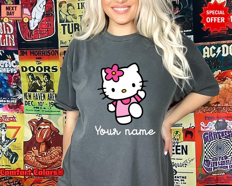May include: A charcoal gray t-shirt featuring a cartoon image of Hello Kitty wearing a pink dress and a flower. The text "Your name" is printed below the character in a cursive font. The shirt is part of a Comfort Colors collection.