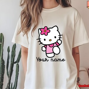 May include: Cream-colored t-shirt featuring a cartoon cat character with a pink flower, wearing a pink outfit. The text "Your name" is written in a script font below the character. The shirt is a Comfort Colors brand.