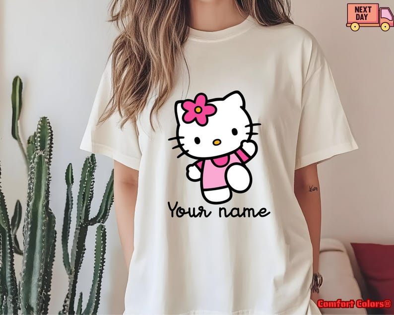 May include: Cream-colored t-shirt featuring a cartoon cat character with a pink flower, wearing a pink outfit. The text "Your name" is written in a script font below the character. The shirt is a Comfort Colors brand.