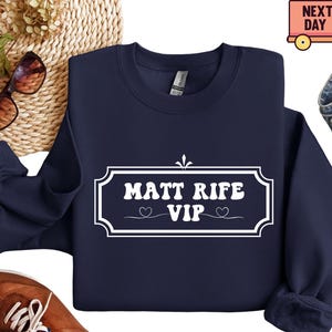 May include: A navy blue sweatshirt with the text "MATT RIFE VIP" in a white, decorative frame. The design includes two small heart symbols. The sweatshirt is displayed on a woven surface with sunglasses and a shoe.