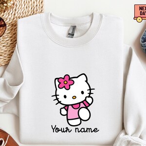 Puede incluir: Sudadera blanca con un personaje de gato de dibujos animados con una flor y un vestido rosas. El gato est&aacute; saludando. Debajo del personaje est&aacute; el texto "Your name" en una fuente negra. La sudadera tiene cuello redondo y mangas largas.