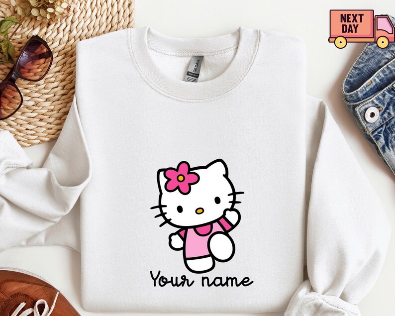 Puede incluir: Sudadera blanca con un personaje de gato de dibujos animados con una flor y un vestido rosas. El gato est&aacute; saludando. Debajo del personaje est&aacute; el texto "Your name" en una fuente negra. La sudadera tiene cuello redondo y mangas largas.