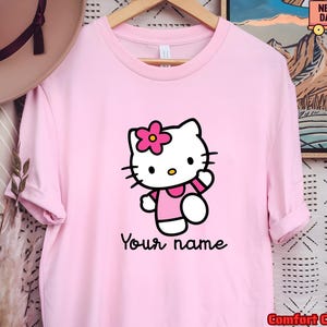 May include: A light pink t-shirt featuring a cartoon cat character with a pink flower on its head. The cat is wearing a pink dress and waving. The text "Your name" is written below the character.