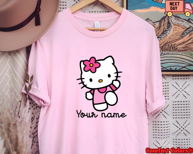 May include: A light pink t-shirt featuring a cartoon cat character with a pink flower on its head. The cat is wearing a pink dress and waving. The text "Your name" is written below the character.