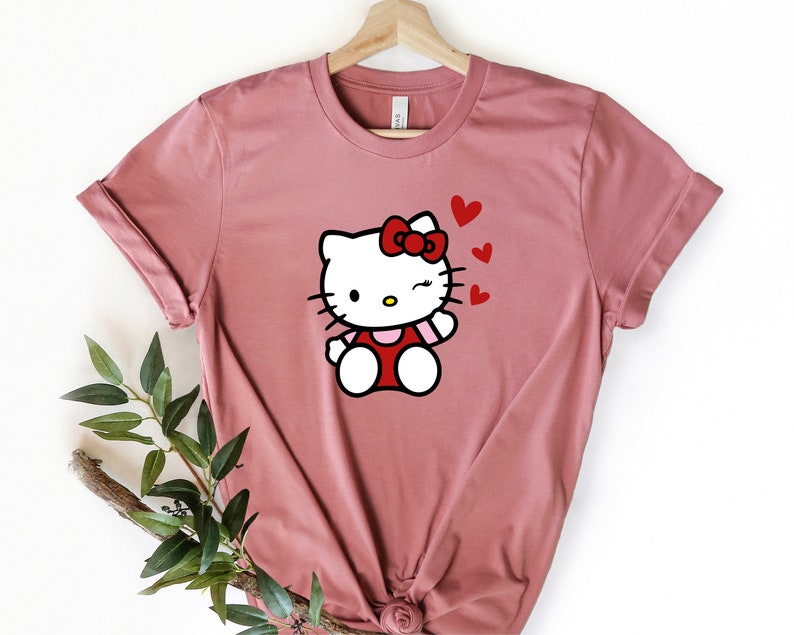 Op de afbeelding: Een roze T-shirt met een witte Hello Kitty-afbeelding die een rode jurk en een rode strik draagt. Hello Kitty knippert met haar oog en heeft rode hartjes om haar heen.