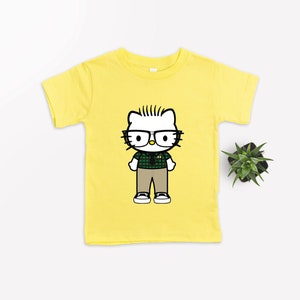 Puede incluir: Camiseta amarilla de manga corta con una imagen de dibujos animados de Hello Kitty con gafas y una camisa a cuadros.