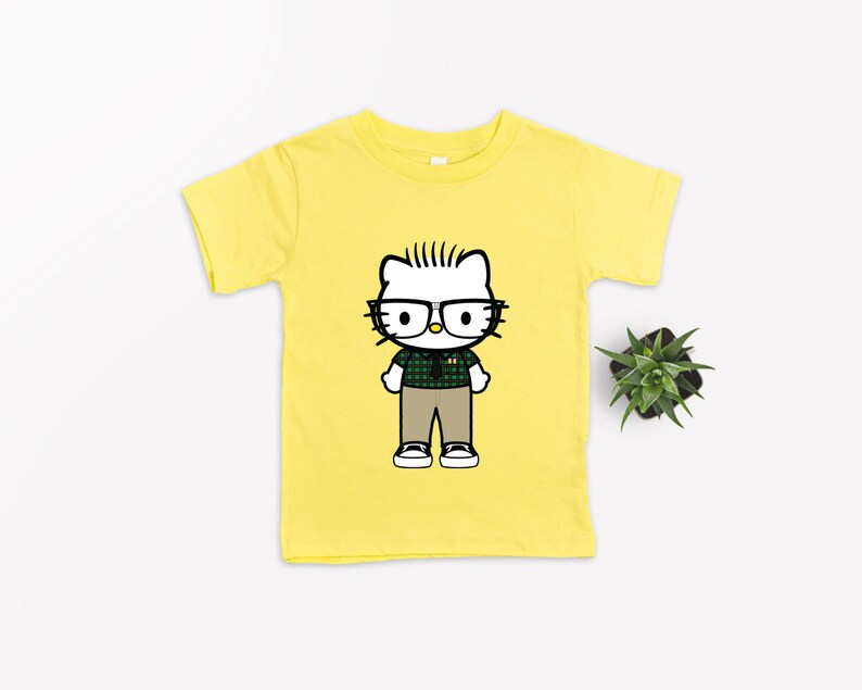 Puede incluir: Camiseta amarilla de manga corta con una imagen de dibujos animados de Hello Kitty con gafas y una camisa a cuadros.