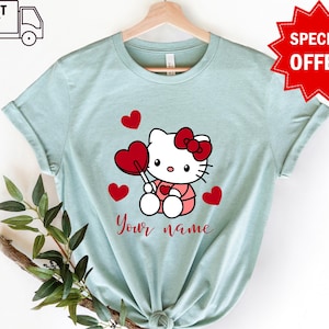 May include: Light blue t-shirt with a cartoon image of Hello Kitty holding a red heart lollipop. There are red hearts around Hello Kitty and the text "Your name" is printed below her.