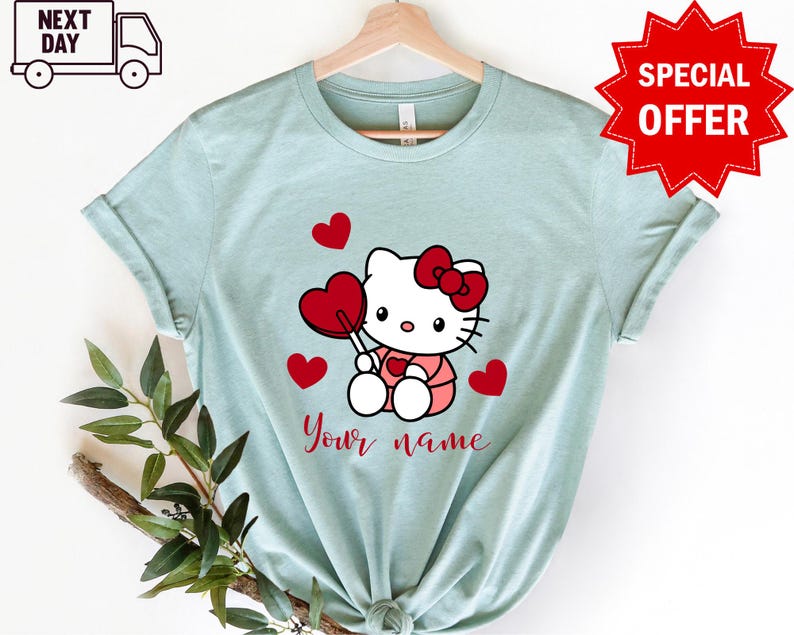 May include: Light blue t-shirt with a cartoon image of Hello Kitty holding a red heart lollipop. There are red hearts around Hello Kitty and the text "Your name" is printed below her.