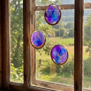 Könnte beinhalten: Drei dekorative Glas-Suncatcher mit einem goldfarbenen Metallrahmen. Das Glas weist wirbelnde Blau- und Lilatöne auf. Die Suncatcher hängen vor einem Holzfenster mit Blick auf eine grüne Landschaft.