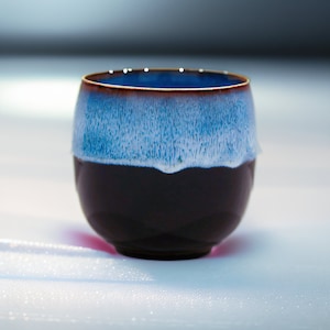 May include: A dark brown ceramic teacup with a light blue and white glaze. The cup has a rounded shape and a wide rim.