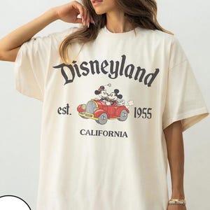 K&ouml;nnte beinhalten: Elfenbeinfarbenes T-Shirt mit dem Disneyland-Logo und einer Grafik von Mickey und Minnie Mouse in einem roten Auto. Der Text "est. 1955 CALIFORNIA" ist in Schwarz aufgedruckt. Das T-Shirt hat kurze &Auml;rmel und eine lockere Passform.