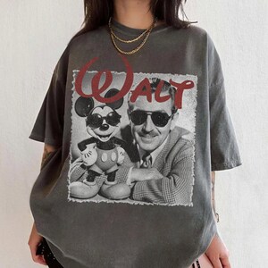 May include: Gray t-shirt featuring a black and white image of Mickey Mouse and Walt Disney with the word "WALT" in red. The shirt has short sleeves and a relaxed fit, suitable for casual wear.