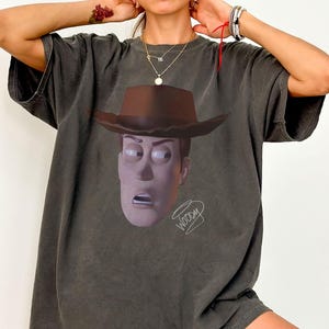 May include: A charcoal grey short-sleeve t-shirt featuring a graphic print of a cowboy character wearing a brown hat. The character has a wide-eyed, surprised expression. Below the character, the word Woody is written in a stylised white script.